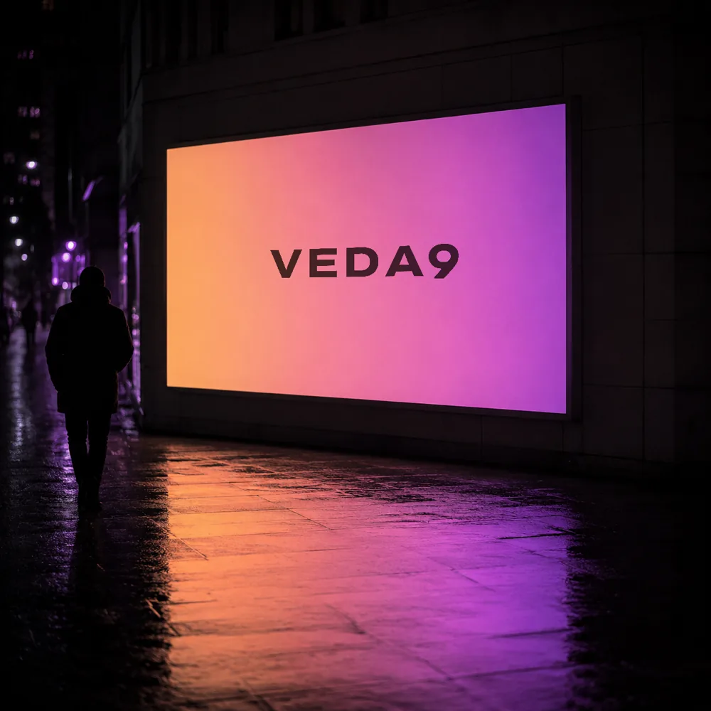 A lit advertising display on a wet street at night carrying the VEDA9 wordmark, a lone figure walking past it.