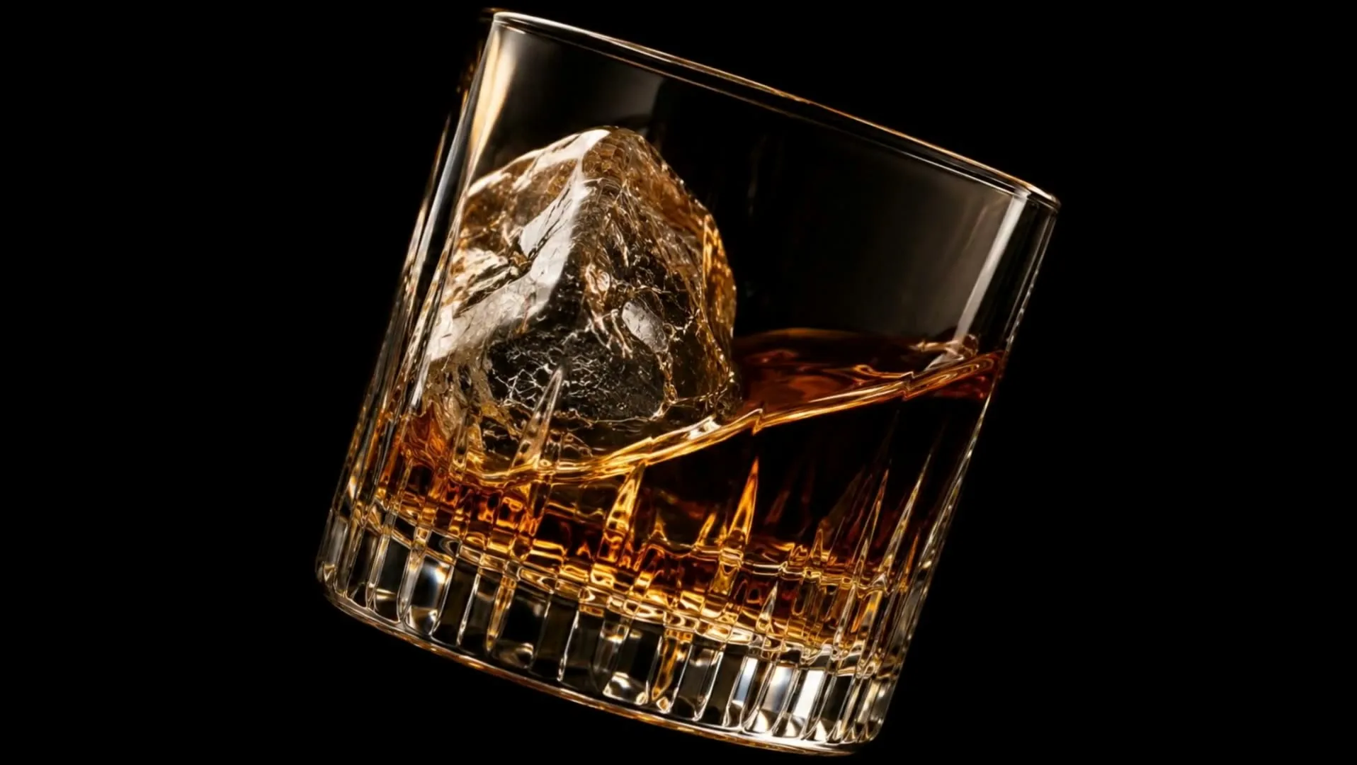 Extreme macro of a rocks glass tilted mid-swirl, whisky riding up one wall against a large clear ice cube, everything around it black.